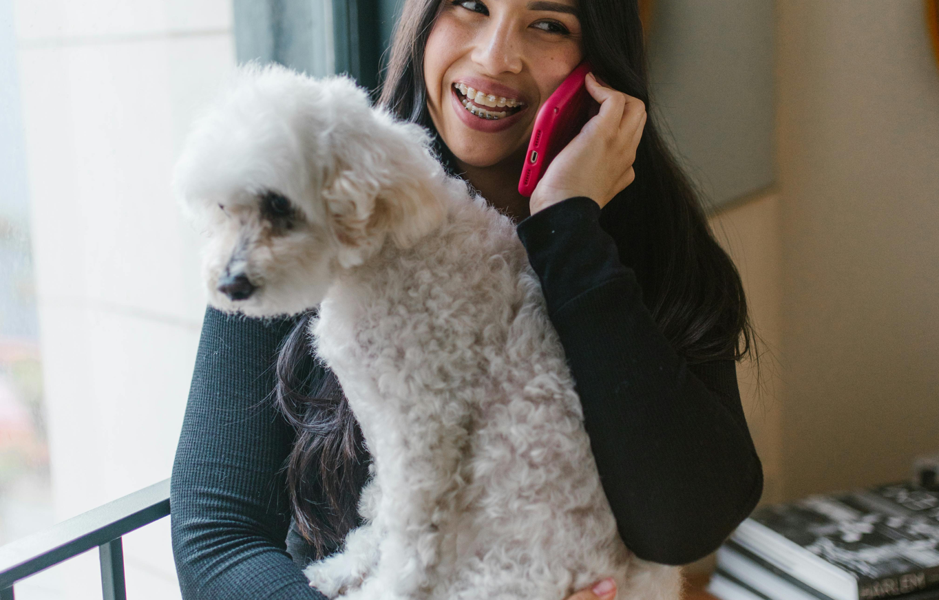 Woman holding white fluffy dog while talking on the phone indoors.